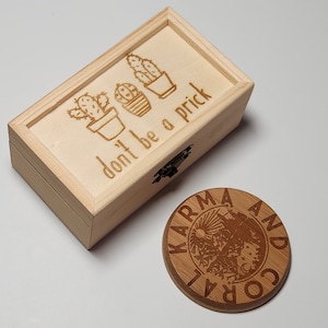 May include: A wooden box with a hinged lid and a small wooden circle. The box is engraved with three potted cacti and the text "don't be a prick". The circle is engraved with the text "Karma and Coral".