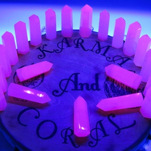 May include: A collection of 18 pink crystal points arranged in a circle on a wooden surface. The surface has the words "Karma And Coral" printed on it.