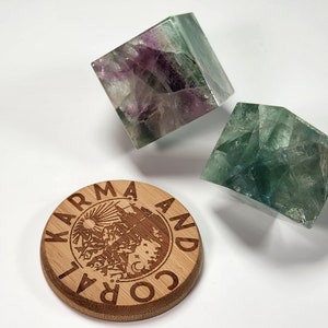 May include: Two green and purple fluorite crystal cubes on a wooden coaster with the text "Karma and Cora" engraved on it.