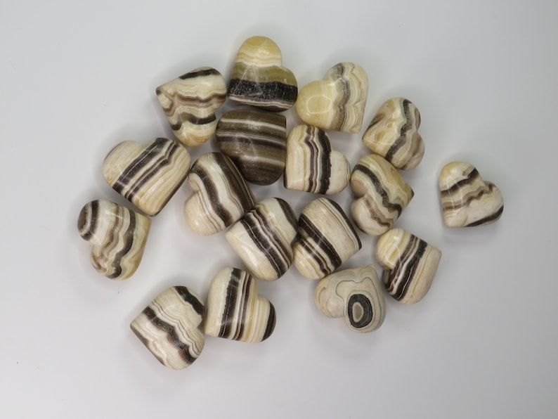 May include: A collection of 15 heart-shaped polished stones with brown and white stripes. The stones are arranged in a random pattern on a white surface.