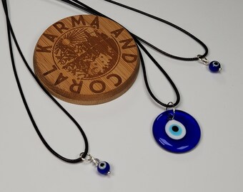 Pode incluir: Três colares de cordão preto com pingentes de olho turco azul. Os pingentes são de tamanhos diferentes, um é uma pequena conta, outro é um pingente redondo de tamanho médio e o maior é um pingente redondo com um design de olho branco e azul. Os colares são exibidos em uma superfície de madeira com as palavras "Karma and Cora" gravadas nela.