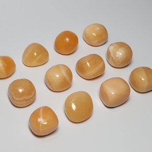 May include: A collection of 12 smooth, polished, yellow-orange carnelian tumbled stones. The stones are various sizes and shapes, with some having white streaks.