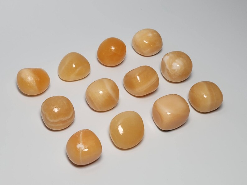 May include: A collection of 12 smooth, polished, yellow-orange carnelian tumbled stones. The stones are various sizes and shapes, with some having white streaks.