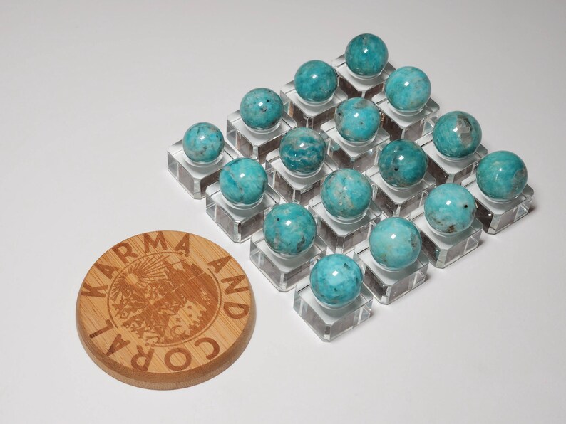 May include: A collection of 20 turquoise blue spheres arranged in a grid pattern on clear acrylic stands. The spheres are all approximately the same size and have a smooth, polished surface. The spheres are displayed on a white surface. A wooden coaster with the text "Karma and Coral" is in the lower left corner of the image.