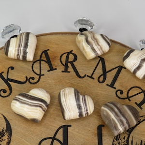 May include: A wooden board with the word "KARMA" written on it. There are eight heart-shaped stones with brown and white stripes on the board. The stones are attached to the board with clear crystal knobs.