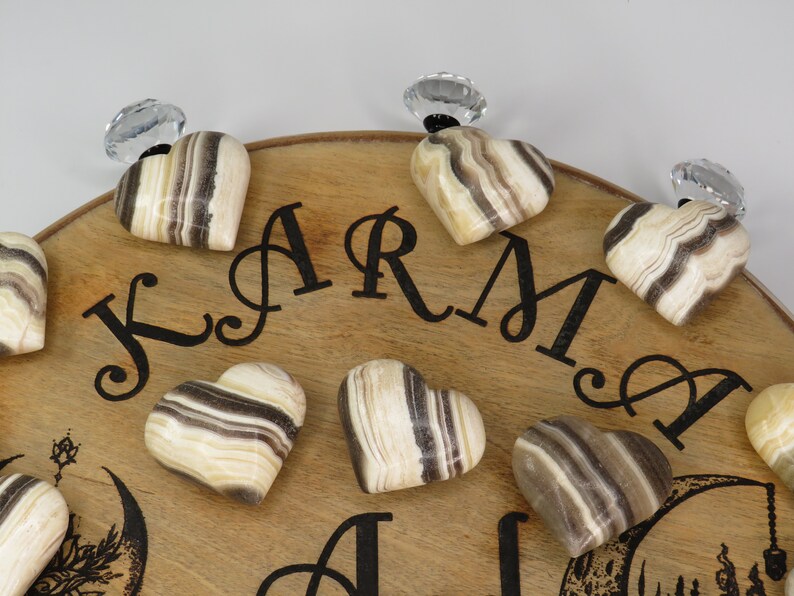May include: A wooden board with the word "KARMA" written on it. There are eight heart-shaped stones with brown and white stripes on the board. The stones are attached to the board with clear crystal knobs.
