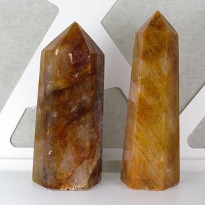 May include: Two golden brown quartz crystal points, also known as citrine, are displayed on a white surface. The crystals are both tall and slender, with a pointed tip and a flat base.