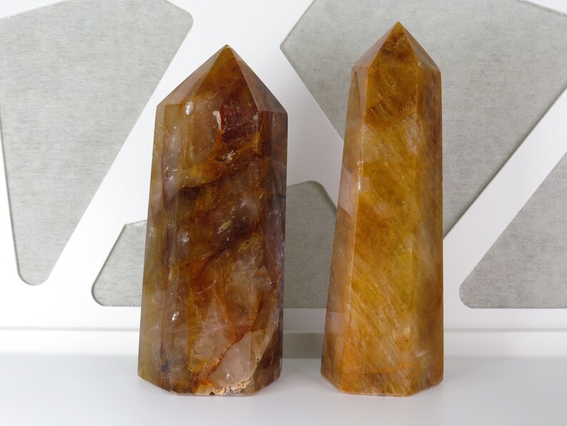 May include: Two golden brown quartz crystal points, also known as citrine, are displayed on a white surface. The crystals are both tall and slender, with a pointed tip and a flat base.