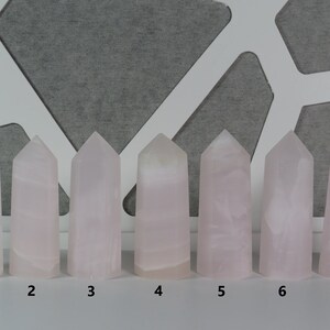 May include: Seven pink crystal points, numbered 1 through 7, arranged in a row on a white surface. The crystals are all roughly the same size and shape, with a pointed top and a flat base.