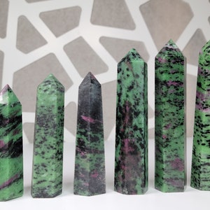 May include: Seven green, black, and pink gemstone points arranged in a row on a white surface. The points are all different sizes and shapes, but they are all polished and have a smooth finish.