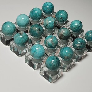 May include: A collection of 18 teal green amazonite spheres displayed on clear acrylic stands. The spheres are polished and have a smooth, glassy finish.