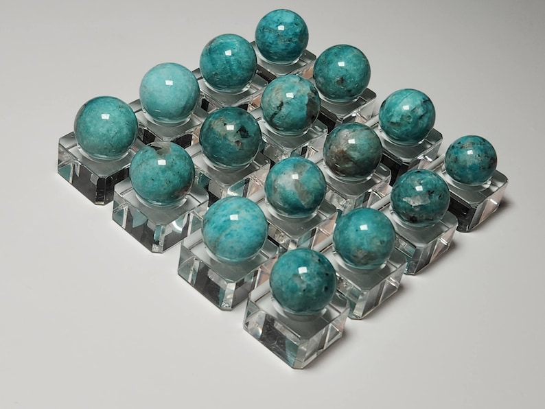 May include: A collection of 18 teal green amazonite spheres displayed on clear acrylic stands. The spheres are polished and have a smooth, glassy finish.