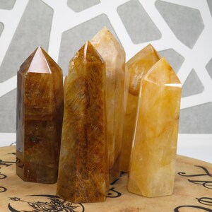 May include: Five golden citrine crystal points arranged on a wooden surface. The points are various sizes and shapes, with some having a more translucent appearance than others.