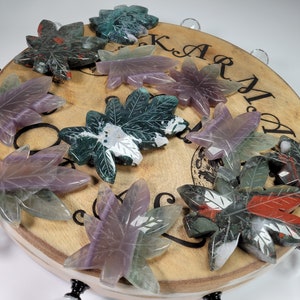 May include: A collection of ten green, purple, and red carved stone maple leaves on a wooden base. The leaves are arranged in a circle around the base, with the text "Karma" inscribed on the base.