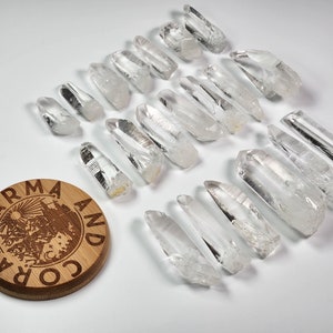 May include: A collection of 20 clear quartz crystal points, also known as rock crystals, arranged on a white surface. The crystals are various sizes and shapes, with some having a slight smoky tint. A small wooden coaster with the text "Karma and Coral" is in the lower left corner of the image.