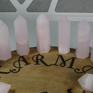 May include: A set of eight pink rose quartz crystal points arranged in a circle on a wooden surface with the word "KARMA" inscribed in black lettering.