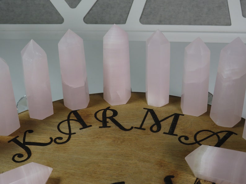 May include: A set of eight pink rose quartz crystal points arranged in a circle on a wooden surface with the word "KARMA" inscribed in black lettering.