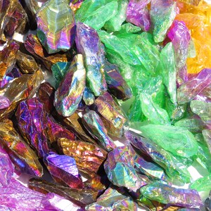 May include: A collection of iridescent crystal points in various colours, including purple, green, gold, and orange. The crystals are arranged in a random pattern on a white surface.