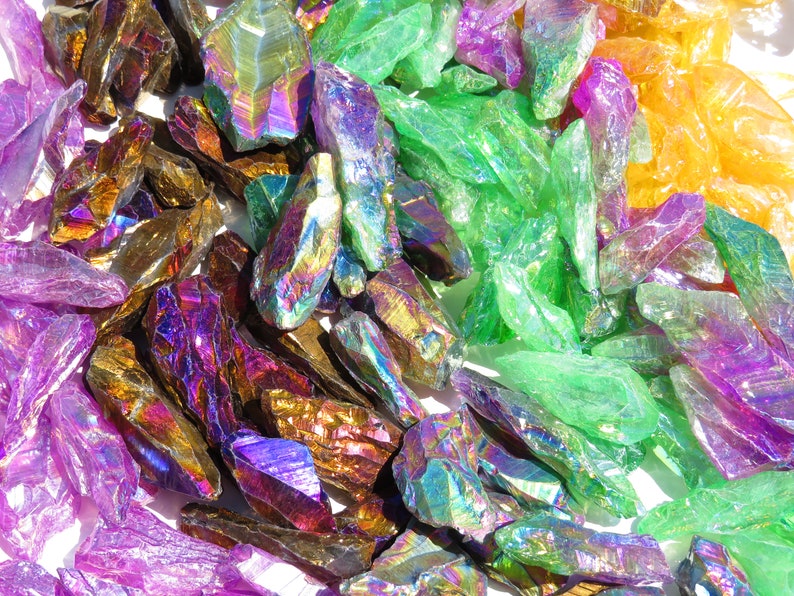 May include: A collection of iridescent crystal points in various colours, including purple, green, gold, and orange. The crystals are arranged in a random pattern on a white surface.