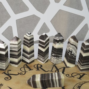 May include: A set of 12 black and white striped obsidian crystal points arranged in a semi-circle on a wooden board with the word "KARMA" inscribed on it.