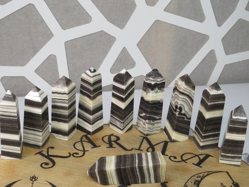 May include: A set of 12 black and white striped obsidian crystal points arranged in a semi-circle on a wooden board with the word "KARMA" inscribed on it.