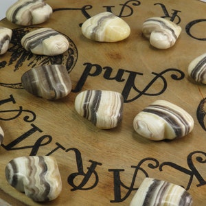 May include: A collection of 11 heart-shaped striped agate stones in shades of brown, white, and cream. The stones are arranged on a wooden surface with black lettering.