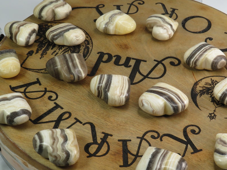 May include: A collection of 11 heart-shaped striped agate stones in shades of brown, white, and cream. The stones are arranged on a wooden surface with black lettering.