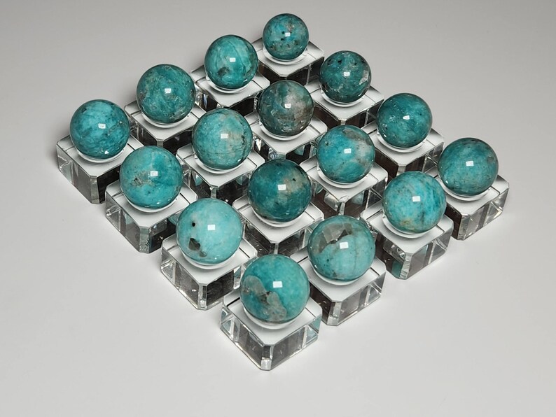 May include: Twenty teal green spheres of amazonite, each displayed on a clear glass pedestal. The spheres are arranged in a grid pattern.