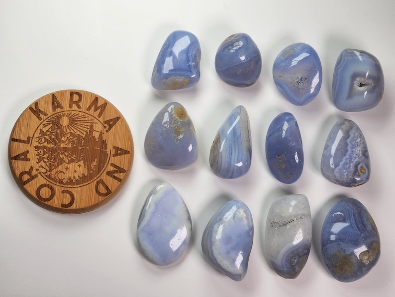 以下が含まれることがあります： 白とグレーの縞模様が入った、滑らかで磨かれた青い瑪瑙の石が12個。石は白い表面に円形に並べられています。左上の画像には、「Karma and Co.」という文字が刻まれた丸い木製のディスクがあります。