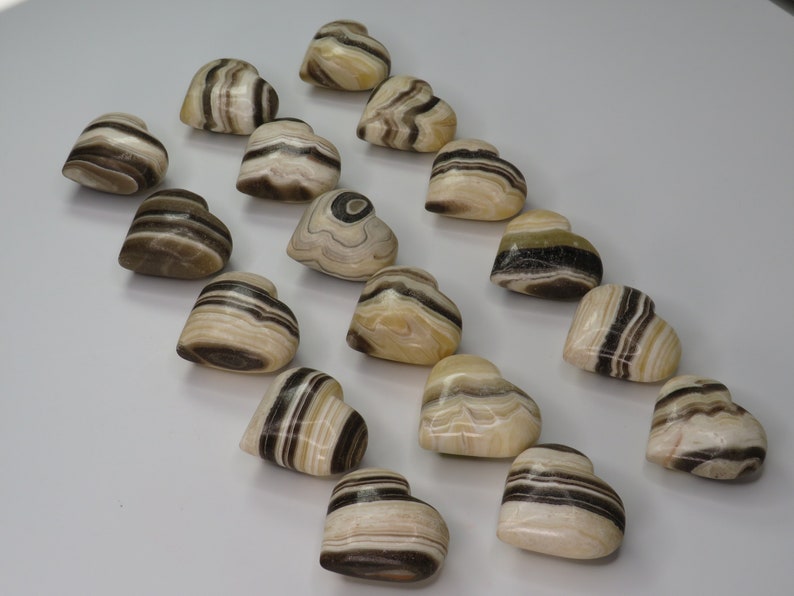 May include: A collection of 18 heart-shaped polished stones with brown and white stripes. The stones are arranged in a row on a white surface.
