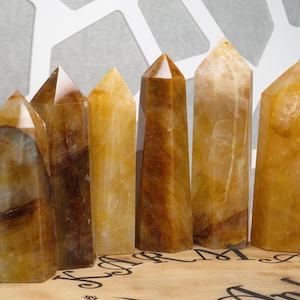 May include: Six yellow and brown crystal points, also known as citrine, arranged in a row on a wooden surface. The crystals are polished and have a smooth, glassy finish.