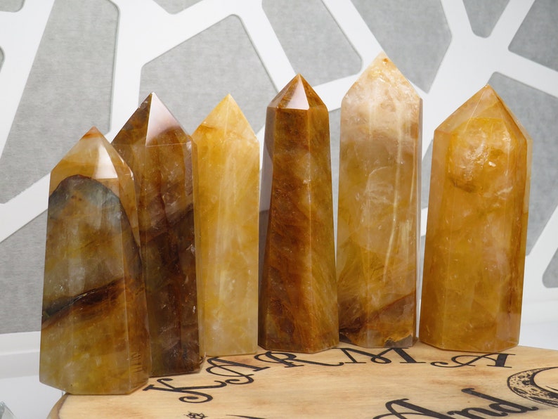 May include: Six yellow and brown crystal points, also known as citrine, arranged in a row on a wooden surface. The crystals are polished and have a smooth, glassy finish.