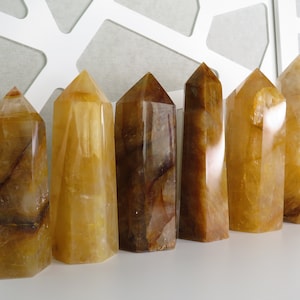 May include: Seven yellow and brown crystal points, also known as citrine, arranged in a row on a white surface. The crystals are all different sizes and shapes, but they are all polished and have a smooth, glassy finish.