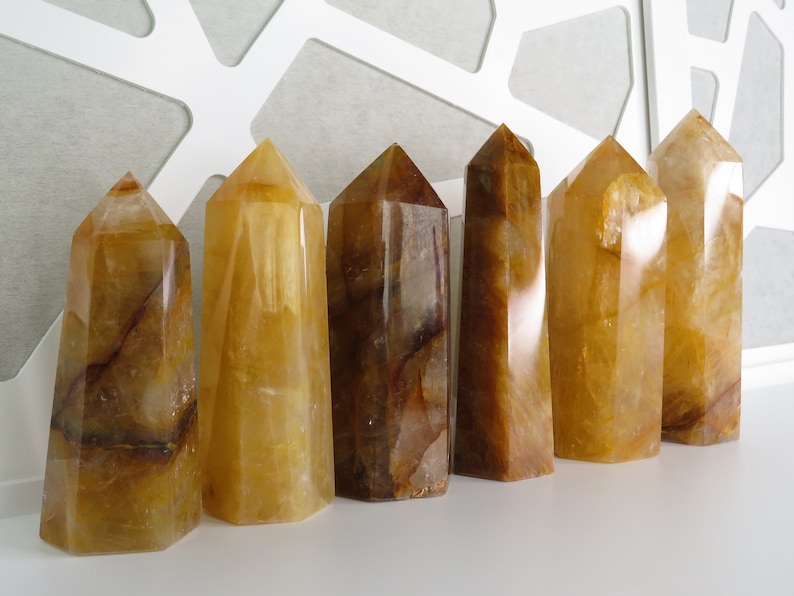 May include: Seven yellow and brown crystal points, also known as citrine, arranged in a row on a white surface. The crystals are all different sizes and shapes, but they are all polished and have a smooth, glassy finish.
