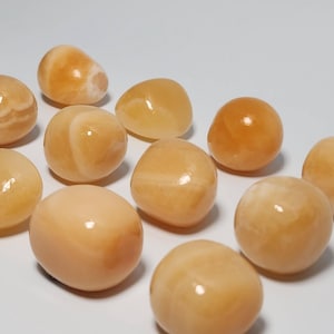 May include: A collection of 11 smooth, polished, orange-colored tumbled stones. The stones are various sizes and shapes, with some having a lighter, almost white, color.