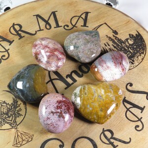 May include: Six heart-shaped polished stones in various shades of red, brown, yellow, and green on a wooden surface with the words "Karma and Soul" engraved on it.