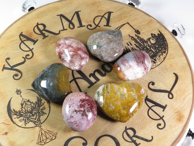 May include: Six heart-shaped polished stones in various shades of red, brown, yellow, and green on a wooden surface with the words "Karma and Soul" engraved on it.