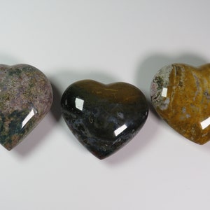 May include: Three heart-shaped polished stones with different patterns and colours. The stones are a light purple with green and brown flecks, a dark green with brown and white flecks, and a yellow with brown and white flecks.