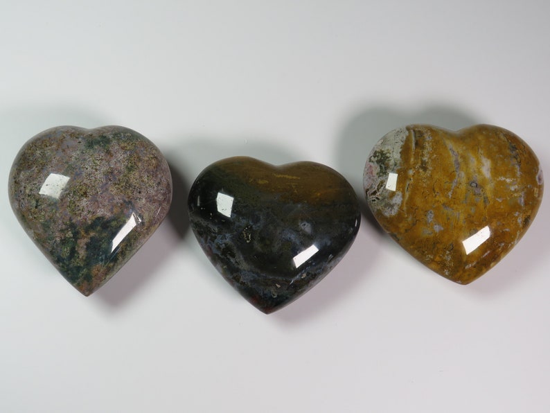 May include: Three heart-shaped polished stones with different patterns and colours. The stones are a light purple with green and brown flecks, a dark green with brown and white flecks, and a yellow with brown and white flecks.