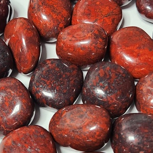 May include: A collection of polished red jasper stones with dark red and black patterns. The stones have a smooth, glossy finish and vary in size and shape. The overall color palette is a deep red with darker, mottled inclusions, creating a natural, earthy aesthetic.