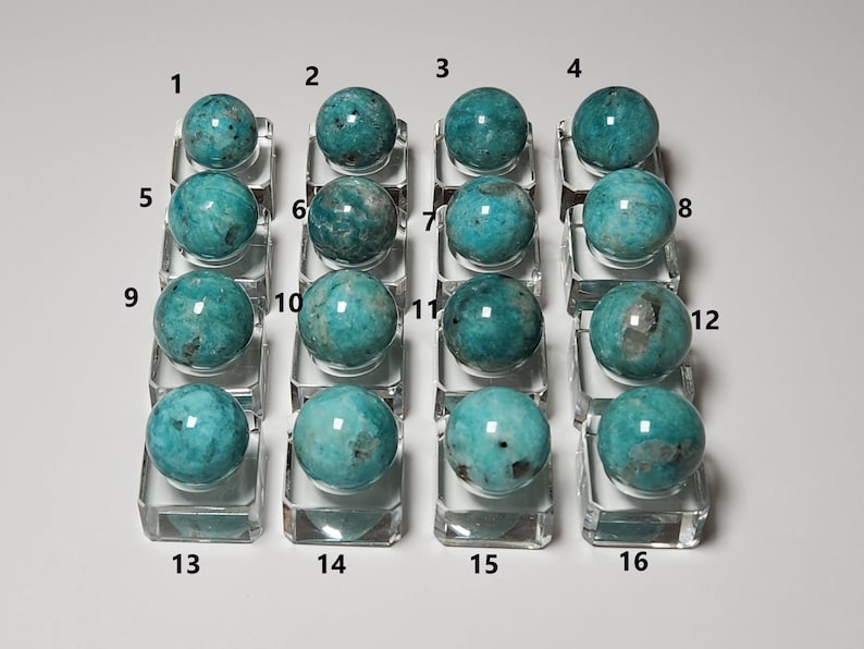 May include: A collection of 16 teal green amazonite spheres displayed on clear glass bases. The spheres are numbered 1 through 16.