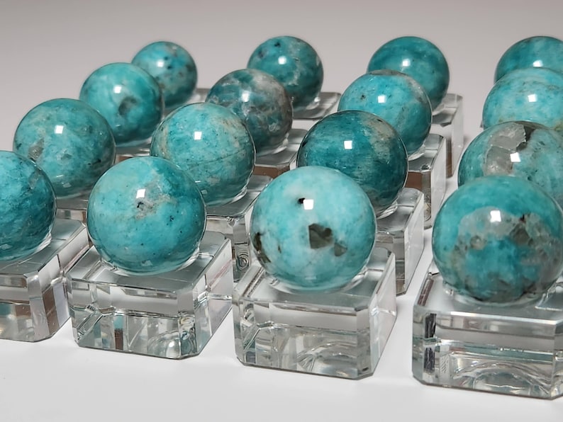 May include: A collection of 15 teal green amazonite spheres on clear glass display stands. The spheres are polished and have a natural, cloudy appearance.