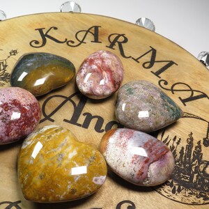 May include: Six heart-shaped polished stones with various patterns of red, pink, white, brown, and yellow. The stones are arranged on a wooden surface with the words "Karma" and "Angel" engraved on it.