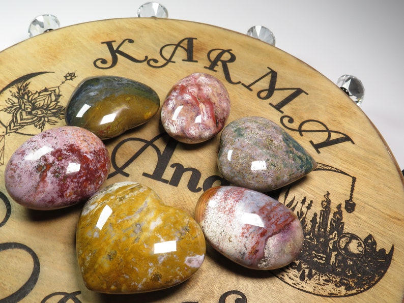 May include: Six heart-shaped polished stones with various patterns of red, pink, white, brown, and yellow. The stones are arranged on a wooden surface with the words "Karma" and "Angel" engraved on it.