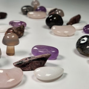 May include: A collection of polished stones and crystals in various shapes and colors, including purple, pink, gray, and brown. Some of the stones are shaped like mushrooms.