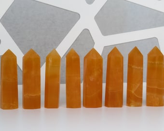 May include: Ten orange calcite crystal points arranged in a row on a white surface. The points are all approximately the same size and shape.