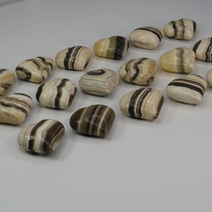 May include: A collection of 17 heart-shaped polished stones with brown and white stripes. The stones are arranged in a row on a white surface.