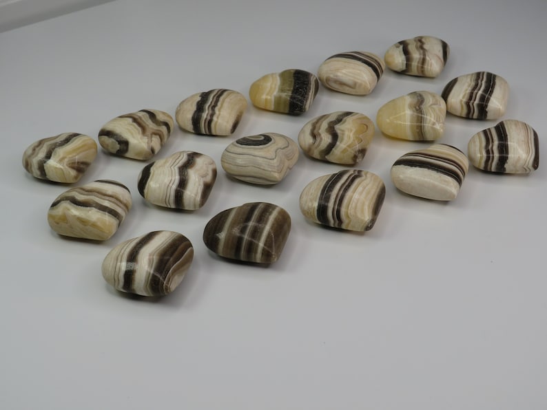 May include: A collection of 17 heart-shaped polished stones with brown and white stripes. The stones are arranged in a row on a white surface.