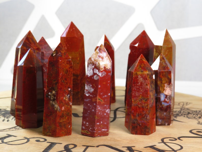 May include: A group of ten red, brown, and white jasper crystal points. The points are arranged in a circle on a wooden surface.
