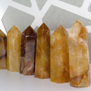 May include: Seven golden citrine crystal points, varying in size and shade, arranged in a row on a white surface. The crystals are translucent and have a warm, golden hue.